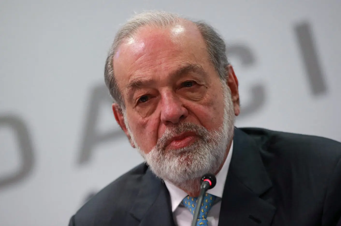 Carlos Slim, com cabelos grisalhos e barba, está sentado e falando em um microfone durante uma conferência. Ele veste um terno escuro e uma camisa azul clara com uma gravata que tem pequenos detalhes em amarelo. O fundo é branco com letras parcialmente visíveis e desfocadas