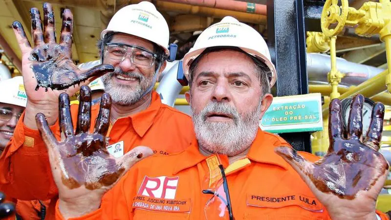 O presidente Luiz Inácio Lula da Silva, em primeiro plano, vestindo um roupão laranja. Trata-se do uniforme da Petrobras. Na imagem, Lula aparece com barba grande e branca. Ele mostra a palma das duas mãos, pretas por causa do óleo da Petrobras