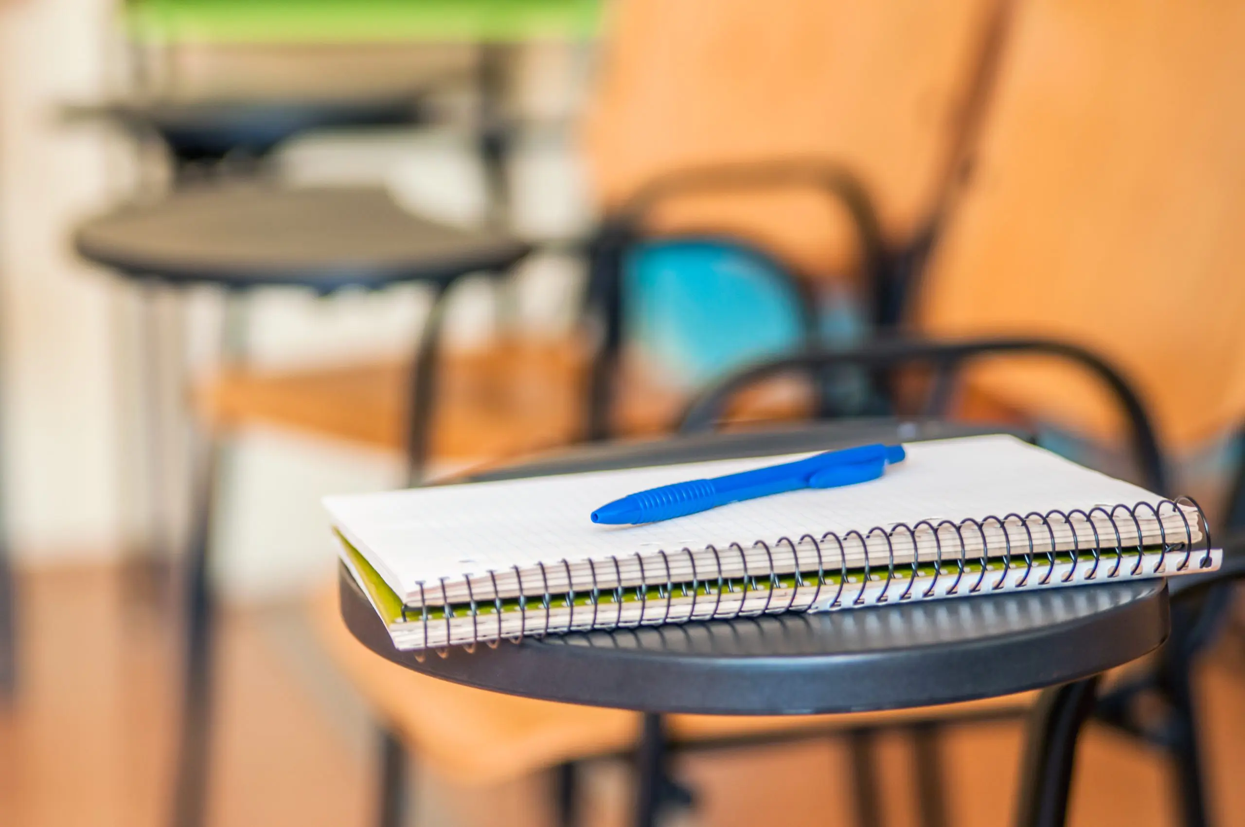 Caderno escolar em cima de uma mesa de estudo preta. As folhas são brancas, e há uma caneta de cor azul em cima do caderno