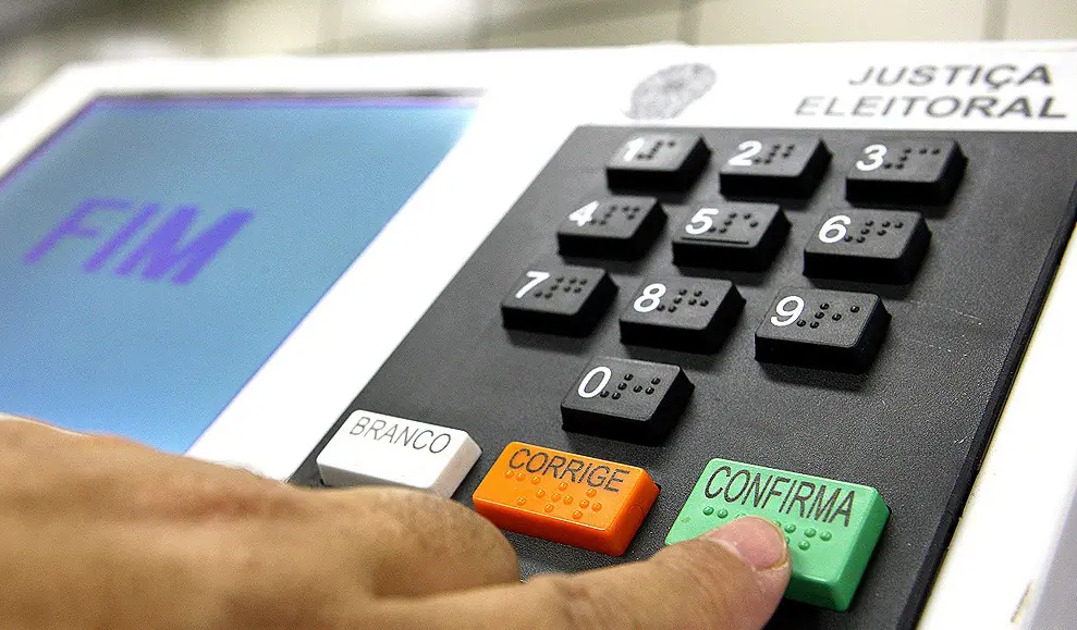 Urna eletrônica brasileira com tela azul exibindo a palavra "FIM" e teclado numérico, o dedo de um eleitor está prestes a pressionar o botão 'CONFIRMA', verde, para finalizar o voto nas eleições municipais