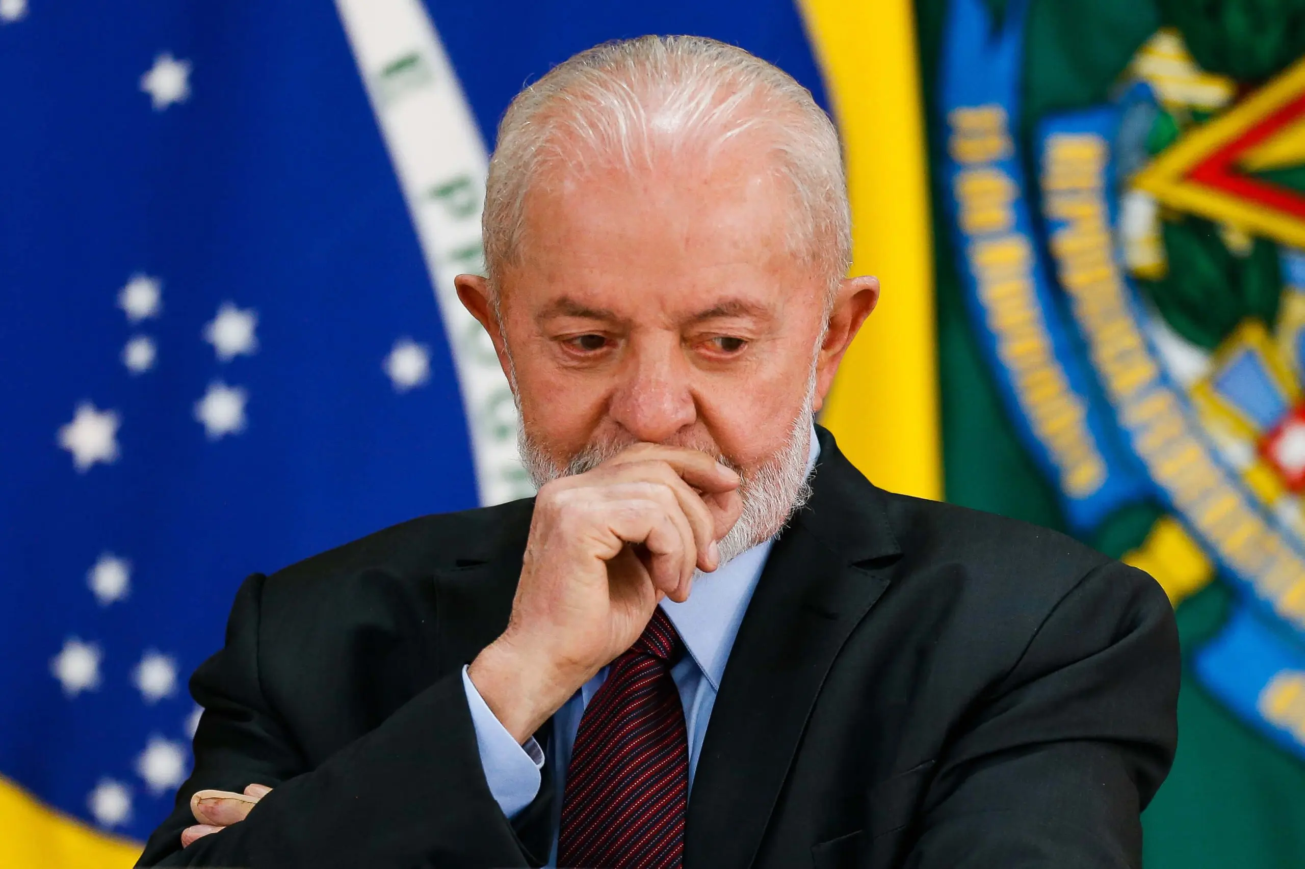 Lula, com a mão direita no rosto; ele está no Palácio do Planalto, em Brasília, usando paletó preto, gravata vinho e camisa azul claro; ele está com semblante de alguém sério, pensativo; atrás dele, há a bandeira do Brasil