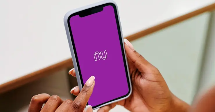 Foto mostra mãos femininas segurando celular. Na tela, aparece o logo do Nubank