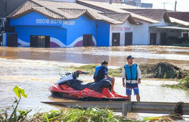 Imagem de bairro alagado no município de Eldorado do Sul (RS); homem em pé aparece de colete salva-vida, há um garoto sentado sobre um jet ski em rua alagada