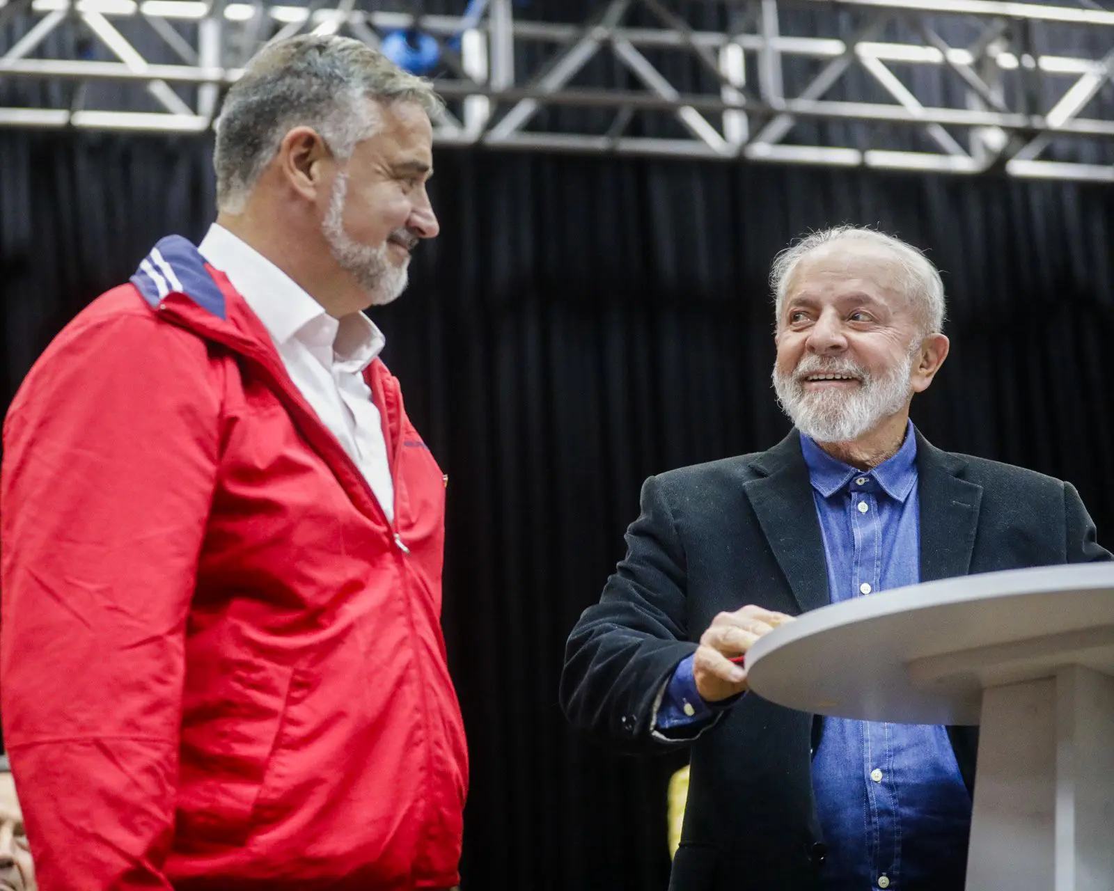 Ministro extraordinário do RS, Paulo Pimenta, e o presidente Lula (PT) estão lado a lado, em frente a um púlpito. Lula usa terno e camisa, e Paulo, roupa de frio vermelha