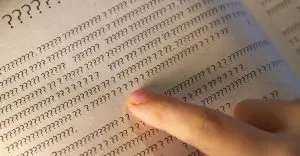 Uma pessoa passa um dedo sobre as linhas do livro, mas em vez de letras, existem apenas pontos de interrogação na página do livro didático