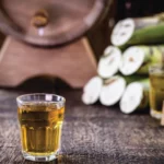 Imagem mostra um copo cheio de cachaça; atrás, há um alambique e cana-de-açúcar, matéria-prima da bebida