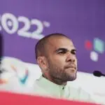 O jogador Daniel Alves, sentado à mesa para uma coletiva de imprensa. Atrás dele, um banner roxo, com o número 2022 estampado