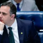 O presidente do Senado, Rodrigo Pacheco, sentado em uma cadeira azul, durante sessão no Congresso Nacional. Ele aparece com a mão no queixo, olhando para o lado direito dele