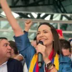 María Corina Machado sorrindo e com um microfone na mão, em meio ao público, com o braço levantado