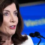 A governadora Nova York, Kathy Hochul. Ela veste camisa branca e está diante de microfones, respondendo a perguntas de jornalistas. Atrás dela, há um banner azul