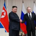 O ditador da Coreia do Norte, Kim-Jong-un, e o presidente da Rússia, Vladimir Putin. Na imagem, ambos aparecem apertando as mãos um do outro. Eles estão em uma sala, com a bandeira dos dois países ao fundo.