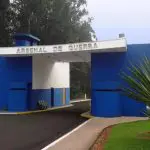 Entrada de um arsenal do Exército com paredes azuis e texto branco, cercada por vegetação, sob céu nublado.