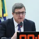 O deputado federal Danilo Forte, em uma sessão no Congresso. Ele está de terno preto, camisa branca e gravata preta. Há uma bandeira do Brasil atrás dele, e um quadro eletrônico à frente.