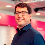 O CEO do iFood, Fabrício Bloisi, postando para foto. Ele está de camisa social azul, óculos, olhando de frente para a Câmera. Ao fundo, uma parede rosa.