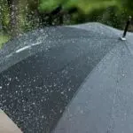 Um guarda-chuva preto, aberto, com água da chuva caindo sobre ele. A previsão do tempo indica chuva