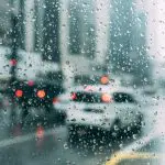 Gotas de água sobre uma lente, oriundas da chuva que caiu sobre a região de algum país. Carros aparecem enfileirados no asfalto, esperando a movimentação do farol