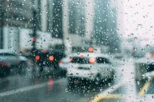 Gotas de água sobre uma lente, oriundas da chuva que caiu sobre a região de algum país. Carros aparecem enfileirados no asfalto, esperando a movimentação do farol