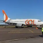 Um avião da companhia aérea Gol, branco com detalhes em laranja. Atrás, um céu aberto. À frente, um homem orientando o aeronauta.
