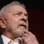 O presidente Luiz Inácio Lula da Silva, com a mão no queixo, olhando para a frente. O fundo da imagem é preto