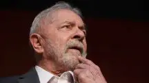 O presidente Luiz Inácio Lula da Silva, com a mão no queixo, olhando para a frente. O fundo da imagem é preto
