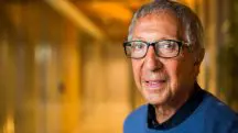 O empresário Abilio Diniz, que faleceu neste domingo, 18. Na imagem, o empresário aparece de camiseta azul, óculos no rosto, olhando para a câmera. Ao fundo, um corredor iluminado em laranja.
