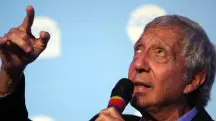 Imagem do empresário Abilio Diniz. Com microfone em mãos, ele olha para cima e aponta para o céu. O fundo da imagem é azul.