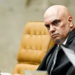 Moraes | O ministro Alexandre de Moraes, com o terno e a túnica preta. Ao fundo, uma cadeira estofada bege, com a parede na cor creme atrás.
