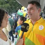 O senador Flávio Bolsonaro, de camiseta amarela com detalhes verdes e laranjas, concede entrevista para uma jornalista