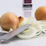 Duas cebolas com casca, em cima de um preto, cercadas por rodelas de cebola cortadas em rodelas. Uma faca está sobre o legume