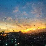 Vista de um pôr do sol visto através de uma janela com gotas de chuva, destacando o clima úmido após uma tempestade