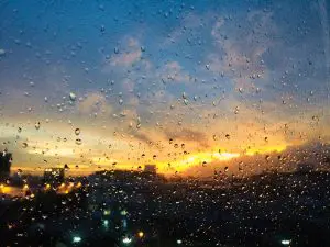 Vista de um pôr do sol visto através de uma janela com gotas de chuva, destacando o clima úmido após uma tempestade