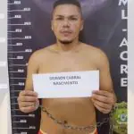 Deibson, integrante do Comando Vermelho, segura um papel com seu nome escrito. Ao fundo, um banner preto da Polícia Civil