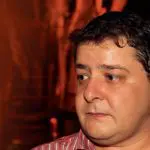 Fabio Luís Lula da Silva, filho do presidente Lula da Silva. Ele aparece olhando para o lado, com camiseta listrada em vermelho e branco. O fundo da imagem é preto e vermelho.