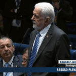 O líder do governo no Senado, Jaques Wagner, em discurso na Casa Alta. Ele está de terno preto, camiseta branca e gravata azul. Ao lado, o senador Omar Azis, sentado, vestido de terno preto