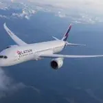 Avião branco da Latam, em voo. Está com as asas curvadas, sobre nuvens, montanhas nevadas ao fundo, sob céu azulado.