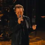 O vocalista Bono Vox, no palco de um show em Las Vegas, pede aplausos a Alexei Navalny, opositor de Putin morto na prisão. O piso do palco é laranja, e o fundo da imagem é preto.
