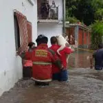 Dois bombeiros, vestidos de vermelho, ajudam a salvar um cachorro durante uma enchente na Bolívia. Na frente dos bombeiros, um homem de camiseta preta. A água, na cor marrom, cobre o trio até a altura da cintura.