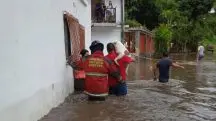 Dois bombeiros, vestidos de vermelho, ajudam a salvar um cachorro durante uma enchente na Bolívia. Na frente dos bombeiros, um homem de camiseta preta. A água, na cor marrom, cobre o trio até a altura da cintura.