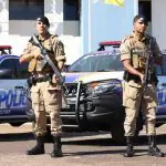 Policiais militares, vestidos de cor bege, seguram armas em frente a dois carros da Polícia Militar