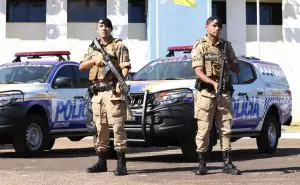 Policiais militares, vestidos de cor bege, seguram armas em frente a dois carros da Polícia Militar
