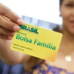 Uma mulher de pele parda segura o cartão de benefício do Bolsa Família, na cor amarela