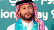 Neymar, de lenço quadriculado na cabeça, nas cores vermelha e branca, com uma faixa preta na cabeça. A roupa é tipicamente árabe. Ele veste um manto branco, com um detalhe azul claro ao redor do pescoço