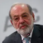 Carlos Slim, com cabelos grisalhos e barba, está sentado e falando em um microfone durante uma conferência. Ele veste um terno escuro e uma camisa azul clara com uma gravata que tem pequenos detalhes em amarelo. O fundo é branco com letras parcialmente visíveis e desfocadas