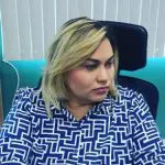 Luciane Farias, a Dama do Tráfico, conhecida como Dama do Tráfico, sentada em uma cadeira preta. Ela tem os cabelos loiros e veste uma camisa azul e branca quadriculada.