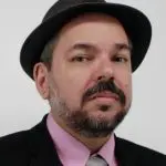 O professor Mario Gazziro, perito da Fórum, vestido de terno preto e camisa rosa. Ele usa chapéu na cabeça. Tem barba grisalha.