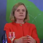 gleisi hoffmann petrobras - Gleisi Hoffmann, com cabelo loiro na altura dos ombros, vestindo um blazer vermelho e uma blusa da mesma cor, está sentada com as mãos cruzadas na frente dela. A expressão é séria e contemplativa. Atrás, há um fundo desfocado com cores verdes e azuis. Há uma garrafa de água azul e uma taça com uma bebida clara à frente da petista