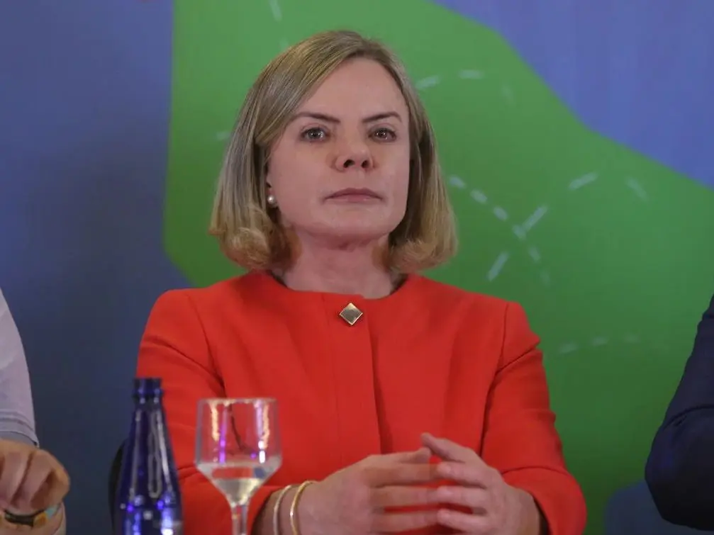 gleisi hoffmann petrobras - Gleisi Hoffmann, com cabelo loiro na altura dos ombros, vestindo um blazer vermelho e uma blusa da mesma cor, está sentada com as mãos cruzadas na frente dela. A expressão é séria e contemplativa. Atrás, há um fundo desfocado com cores verdes e azuis. Há uma garrafa de água azul e uma taça com uma bebida clara à frente da petista