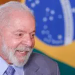 O presidente Luiz Inácio Lula da Silva, sorrindo, com a bandeira do Brasil atrás. Ele usa terno cinza, camisa azul clara e gravata azul com pintas brancas