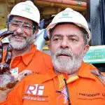 O presidente Luiz Inácio Lula da Silva, em primeiro plano, vestindo um roupão laranja. Trata-se do uniforme da Petrobras. Na imagem, Lula aparece com barba grande e branca. Ele mostra a palma das duas mãos, pretas por causa do óleo da Petrobras
