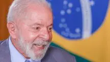 O presidente Luiz Inácio Lula da Silva, sorrindo, com a bandeira do Brasil atrás. Ele usa terno cinza, camisa azul clara e gravata azul com pintas brancas